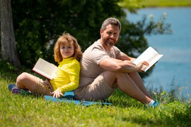 Baba ve oğlu yaz parkında birlikte kitap okuyorlar. Baba oğul parkta piknik yapıyor. Mutluluk aile kavramı. Baba ve oğul gündüz vakti parkta oynuyorlar. Arkadaş canlısı aile.