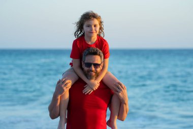 Baba ve oğul gün içinde plajda yürüyorlar. Arkadaş canlısı bir aile. Babalar Günü. Aşk ailesi, baba ve oğlu sahilde deniz doğasında birlikte yürürler. Oğullar babalarının omuzlarına biner.