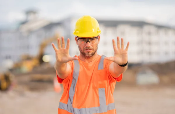 Worker with stop hand gesture. Builder in a hard hat working on a construction project at a site. A builder worker in a helmet near building construction sites. Builder on the job