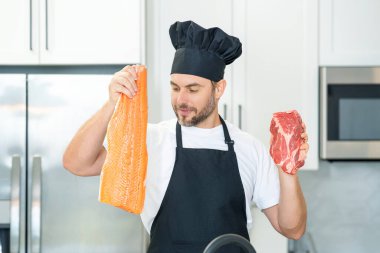 Üniformalı şef mutfakta çiğ et ve balık fileto pişiriyor. Restoran menüsü konsepti. İş üniformalı, siyah önlüklü, şef şapkalı bir aşçı. Balık ve et veya somon ve sığır eti konsepti