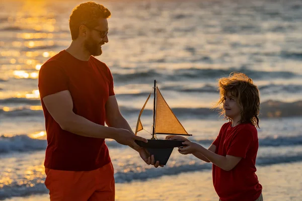 Father and son dream on travel. Father and son enjoying sunset play toys sailing boat. Summer travel, family holidays. Journey trip lifestyle yachting concept. Parenting, fatherhood and fathers day