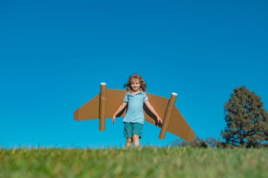 Child boy plays in an astronaut dreams of space. Happy child play with toy plane cardboard wings against blue sky. Kid having fun in summer field outdoor. Portrait of boy with paper wings
