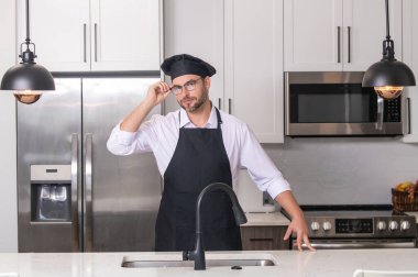 Mutfakta şef üniforması giyen yakışıklı orta yaşlı bir adam. Aşçılık ve aşçılık. Restoran menüsü konsepti. Yemek servisi, yemek pişirme, yemek reklamcılığı. Üniformalı adam mutfakta.