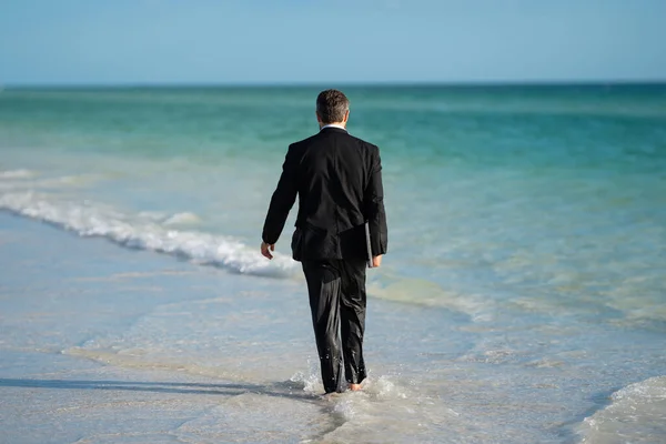 Freelance work, online business. Summer business. Rear view of back businessman in suit in sea water at beach. Funny business man in a business suit with laptop standing in water. Remote work