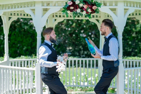 Gay with partner open champagne. Gay couple holding bouquet of flower together during wedding ceremony. Homosexual couple celebrating their wedding. LBGT couple at wedding ceremony