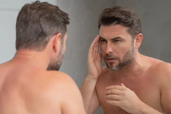 鏡に化粧品の顔クリームを塗る男 顔の治療 裸の肩が肌に触れる男の肖像画 スキンケア製品を持つセクシーな男 男性の顔の犯罪 完璧な肌 朝のルーチン — ストック写真
