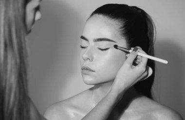 Yüzünde parlak makyaj olan bir modelin yakın plan portresi. Makyöz göz farı sürüyor. Vizecinin eli, genç güzellik modelinin kozmetik ürünlerini boyuyor.