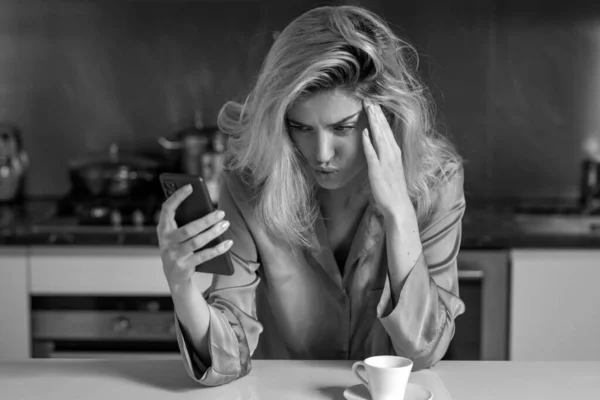Depressed woman in the kitchen in the morning. Sad woman drink coffee and using phone at home in morning. Tired unhappy female housewife chatting smartphone in sexy pajama at kitchen