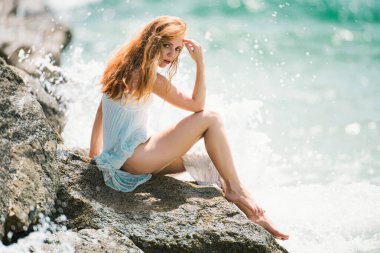 Yaz rüyası. Kayalık sahilde deniz mavisi dalgalara karşı oturan seksi kadın. Kumsalda seksi bir yaz elbisesi giymiş, dirseği dizinde ve eli kafasında şehvetli genç bir kadın.