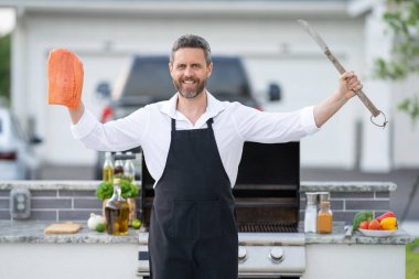 Aşçı önlüğü giymiş erkek aşçı bahçede ızgara somon balığı yapıyor. Barbekü somon filetosu hazırlayan yakışıklı adam. Barbekü ve ızgara. Arka bahçede balık pişiriyorum.