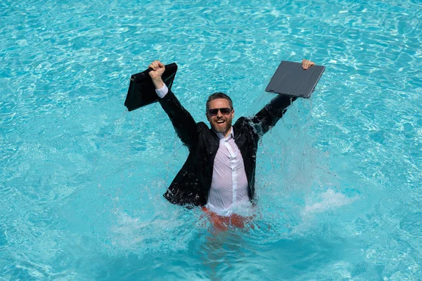 Crazy business. Fun business lifestyle. Handsome excited business man in suit jump with laptop in swimming pool. Funny businessman. Freelance concept, summer travel. Business success