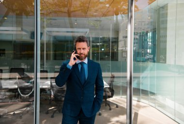 İş adamı telefonda konuşuyor. Modern ofisin yanında dururken cep telefonuyla konuşan neşeli bir işadamı portresi.