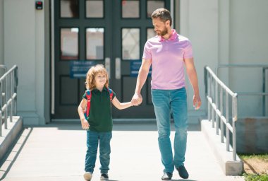Baba oğluna okula kadar eşlik ediyor. Sırt çantalı ilkokul öğrencisinin ebeveyni ve öğrencisi. Ebeveyn çocuğunu okula götürüyor. İlkokul öğrencisi dışarıda sırt çantasıyla çalış.