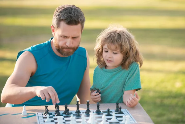 Son laying on grass and playing chess with father. Concentrated kid boy developing chess strategy, playing board game with parent. Games and activities for children