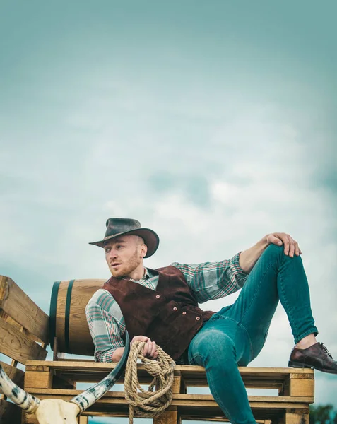 Cowboy farmer man in country side wearing western cowboy hat. American Male model in countryside on farm. Men retro fashion, vintage vogue, brutal male model