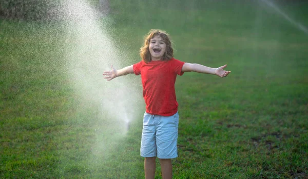 Child play in summer garden. Grass watering. Automatic sprinkler irrigation system in a green park watering lawn. Sprinkler watering. Child gardening concept. Little kid having fun on water spraying