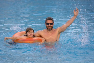 Çocuk havuza su sıçratıyor. Baba ve oğul mavi deniz suyunda yüzüyor ve su sıçratıyorlar. Yaz tatilindeki aile. Mutlu ebeveynler ve çocuklar sahilde dinleniyor. Baba ve çocuk havuz başında oynuyorlar.