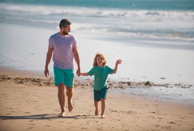 Baba ve çocuk el ele tutuşup birlikte yürüyorlar. Baba oğul yaz plajında yürüyorlar.
