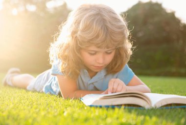 Kid boy reading a book lying on grass. Cute little child in casual clothes reading a book and smiling while lying on grass in park. Smart clever Kids