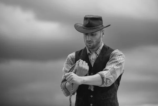 Cowboy farmer man in country side wearing western cowboy hat. American Male model in countryside on farm. Cowboy with lasso rope on sky background