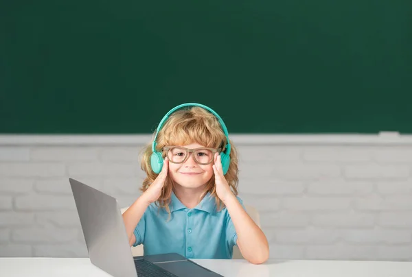 ヘッドフォンの賢い男の子は机に座って 学校でラップトップでオンラインで勉強し 賢い子供はインターネットのレッスンを使ってノートパソコンでイヤホンを身に着けています — ストック写真