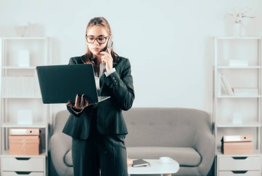 Takım elbiseli ciddi bir iş kadını telefon görüşmesi yapıyor. İş kadını serbest bilgisayar kullanıyor, bilgisayar kullanıyor, bilgisayarda yazıyor, ofiste internette sörf yapıyor.
