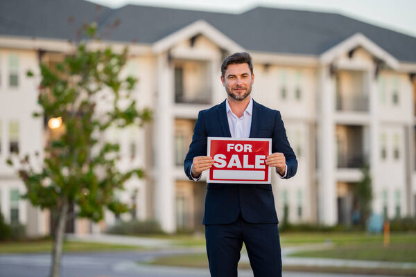 Успешный риэлтор в костюме с вывеской на продажу. Риэлтор перед новым домом. Продавец продает дом, новую квартиру в современном доме