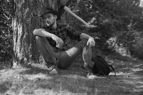木材ジャック労働者の男は森の中に座っている 木立の上に斧を持つルンベルジャック 人間は仕事をしてる 木立の中に斧で立っている木材ジャックの労働者 ハードワーク後の休息 — ストック写真