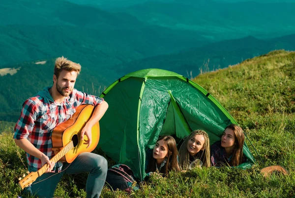 若いです陽気な女性のグループキャンプテント一緒に中に若いですハンサムな男演奏ギター — ストック写真