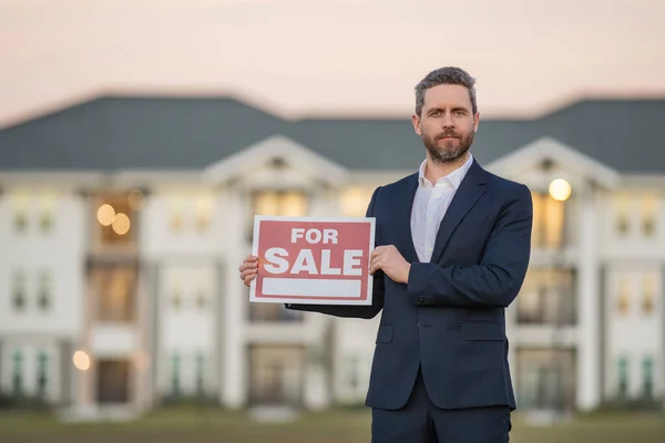 Successful real estate agent in suit with sign for sale. Real estate broker front of new house. Real estate home owner business man salesman sell house, new apartment in modern building