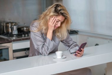 Üzgün kadın sabah kahve içer ve evde telefon kullanır. Mutfakta seksi pijamayla akıllı telefonlardan sohbet eden yorgun bir kadın.