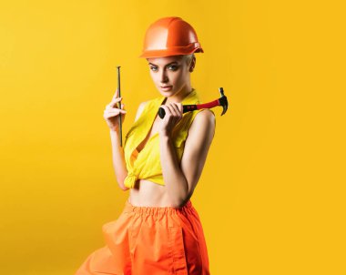 Çekiç kullanmak için kasklı seksi kadın. Sarı arka plandaki atölyede çivi çakan genç bir kadın izole edilmiş. Kadın model bina üzerinde çalışıyor. Her şey düzeltilebilir.