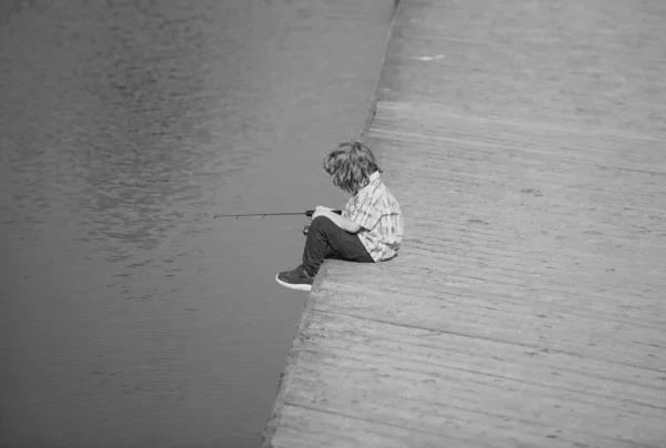 小男孩坐在湖边钓鱼 孩子们的嗜好笑着让孩子在湖上钓鱼 在河边有纺锤的男孩 男孩在防波堤与杆子 复制空间 — 图库照片