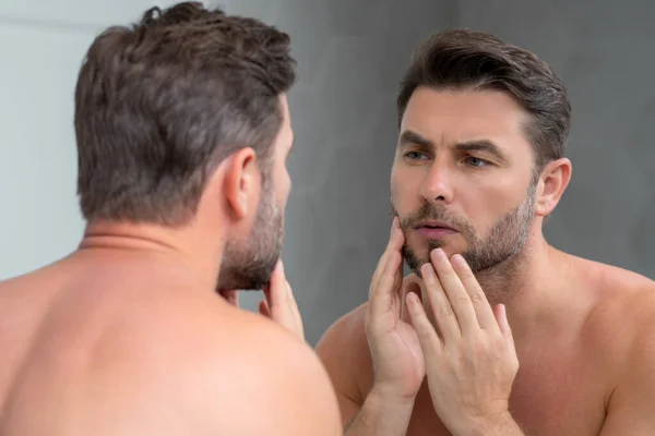 Millennial hispanic man looking in mirror, facial skin and stubble. Male beauty care product. Skincare, home spa. Beauty portrait of a beautiful man. Spa model, moisturizing nourishing creme