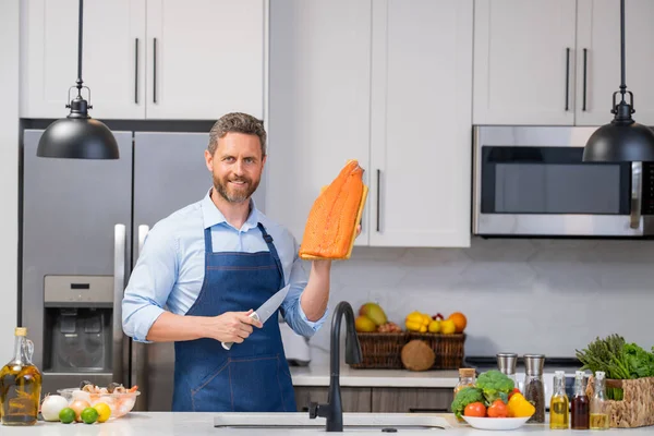 Cook in chef apron with raw fish salmon fillet on kitchen. Chef man in uniform cook apron on kitchen. Handsome man chef in uniform with chef hat cooking raw fish salmon fillet in the kitchen