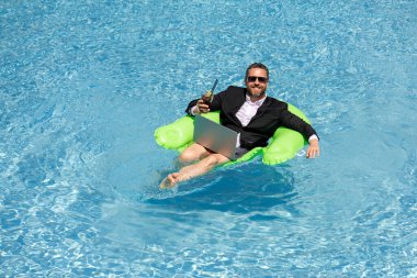 Serbest çalışma, uzaklık, online çalışma, e-çalışma. Yaz işi. Takım elbiseli bir iş adamı yaz kokteyli içer ve bilardoda dizüstü bilgisayar kullanır. İş adamı yazın havuz suyunda iş yapmayı hayal eder.