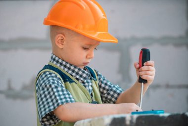 Çocuk tornavidayla cıvata çeviriyor. Tamir aletli bir çocuk tamircisi. İnşaat alanında kasklı çocuk ve içki tulumu var. Küçük işçi mühendisi. Evi onarın.