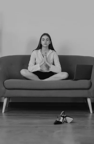 Meditation woman in a modern workplace doing yoga exercise on sofa at workplace in a modern office. Employee feel balance harmony relaxation. Calm accountant girl on sofa in office