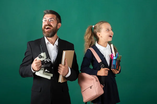 Cute schoolgirl with excited amazed teacher in school. Studio portrait of tutor and young school girl with backpack and textbook on blackboard green background