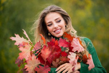 Sonbahar yapraklarını güz doğasında yüzüne tutan mutlu kadın. Sonbahar akçaağaçlı genç bir kadının portresi dışarıda duruyor. Güzel kız parkta sonbahar yaprağıyla oynuyor. Ilık sonbahar. Sonbahar mevsimi modası