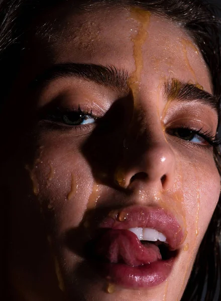Sensual young woman face with honey drop close up. Beautiful girl tongue licking honey lips. Sexy model with honey drip on face
