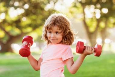 Yeşil parkta dambıllarla egzersiz yapan sevimli küçük çocuk. Dambılı spor bir çocuğun yakın plan portresi. Mutlu çocuk dışarıda egzersiz yapıyor.