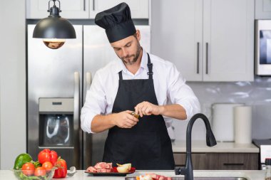 Önlüklü adam ve aşçı şapkalı adam mutfakta yemek pişiriyor. Mutfakta sağlıklı yemek pişiren yakışıklı adam. Adam mutfakta yemek pişiriyor. Taze yemek malzemeleriyle ev menüsü. Modern mutfak iç mimarisi