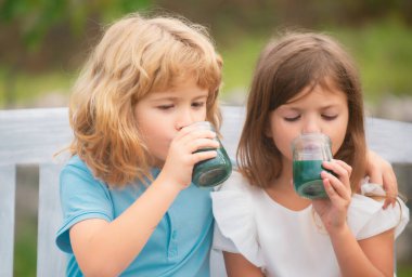 Erkek ve kız kardeş smoothie içer, çocuklar bahar parkında yaz kokteyli içer. Küçük erkek ve kız çocuklar baharın tadını çıkarıyorlar. Çocuklar arka bahçede yürüyor. En iyi arkadaşlar iki çocuk.