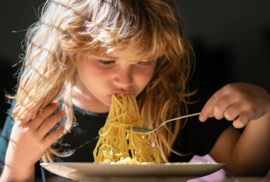 Evde kahvaltı yapan çocuk portresi. Sarı saçlı, tabak kaşığıyla yemek yiyen, yakın plan çocuk yüzlü tatlı bir çocuğun portresi. Evde makarna yiyen şirin bir çocuk.