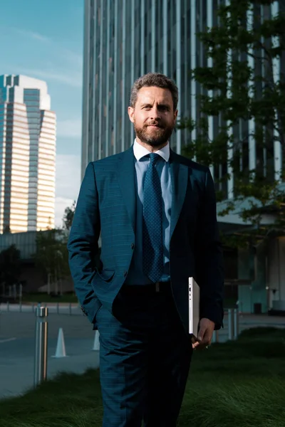 Urban business man. Businessman with laptop standing on the street near business office building. Businessman front of office. Handsome businessman. Business portrait of serious business man outdoor