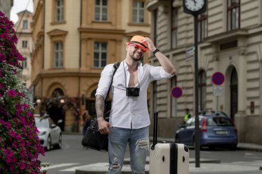 Gezgin, moda turisti. Sıradan giysili şapkalı, bavullu gezgin turist. Seyahat çantası taşıyan bir turist. Elinde bavul olan bir adam şehir caddesinde yürüyor. Hafta sonları seyahat.