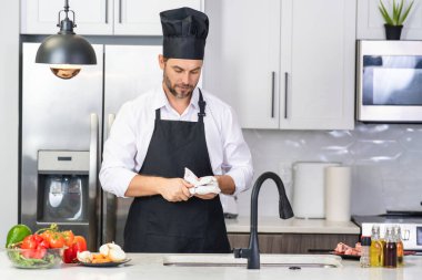 Önlüklü adam ve aşçı şapkalı adam mutfakta yemek pişiriyor. Mutfakta sağlıklı yemek pişiren yakışıklı adam. Adam mutfakta yemek pişiriyor. Taze yemek malzemeleriyle ev menüsü. Modern mutfak iç mimarisi