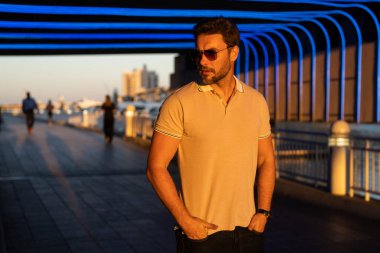 Şehir merkezinde sıradan, yakışıklı bir manken. İyi giyimli, gözlüklü ve polo pozu veren bir adam. Erkek model. Şehirde şık bir adamın portresi. Açık hava moda portresi