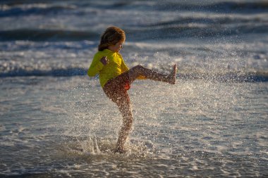 Küçük çocuk suda oynar ve su sıçratır. Çocuk damlalarla eğleniyor. Yazlık çocuk dalgalarda oynuyor, çocuk denizde oynuyor ve su damlaları sıçrıyor. Mayo giymiş bir çocuk yaz boyunca deniz suyuna koşuyor..